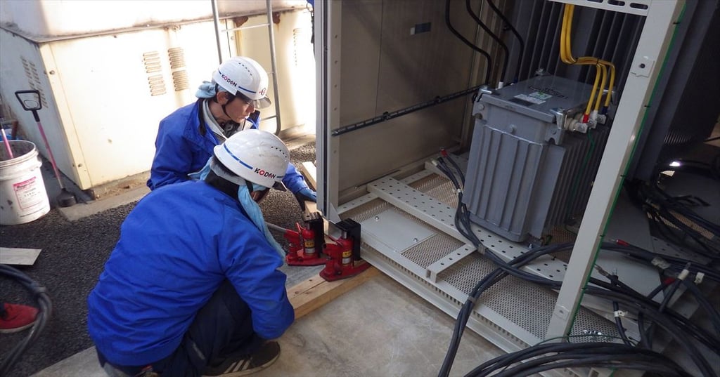新しいキュービクル内に油圧ジャッキで変圧器を据え付ける作業員2名