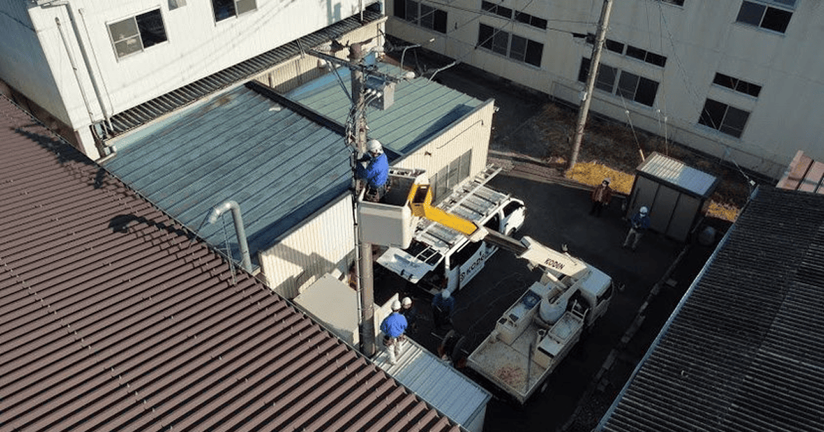 テナントビル裏で高所作業車を使い電柱上の作業を行う恒電社の作業員たち（俯瞰）