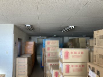 交換前：段ボールが積まれた保管スペースの天井照明